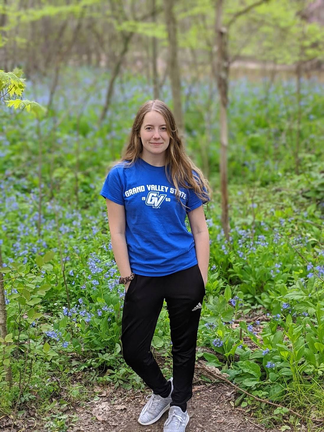 Girl with long blonde hair and blue t shirt standing in the woods with blue flowers in the background